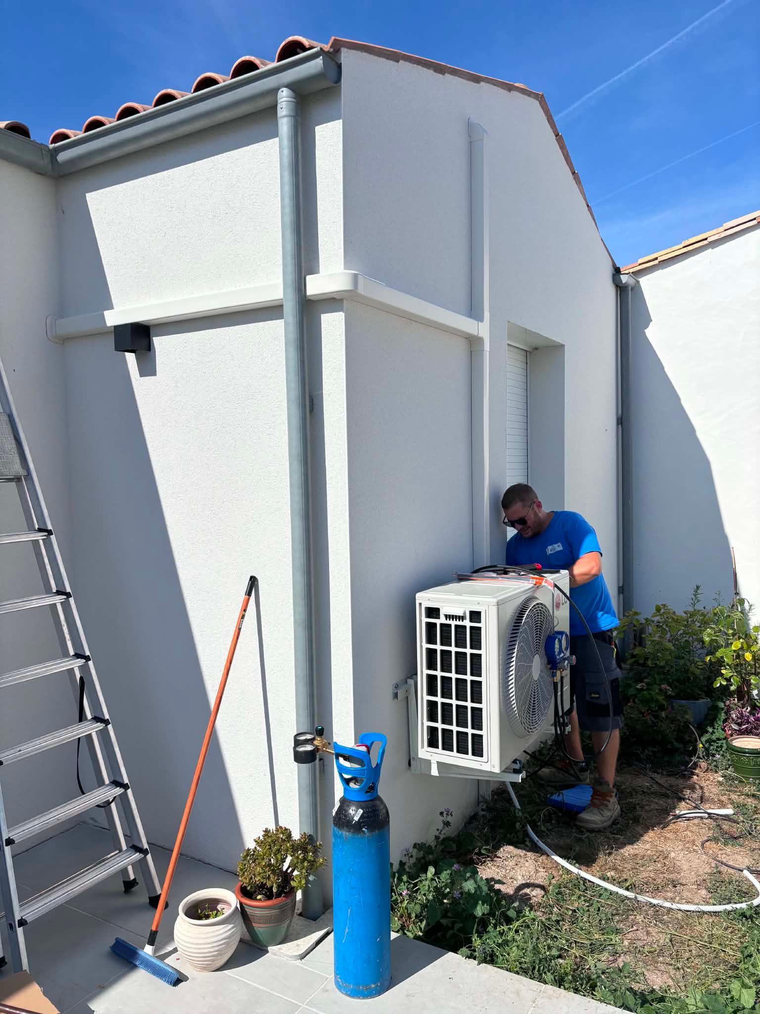 Technicien en train d’entretenir une unité de climatisation extérieure près d’une maison blanche, avec une échelle et un réservoir bleu à proximité.