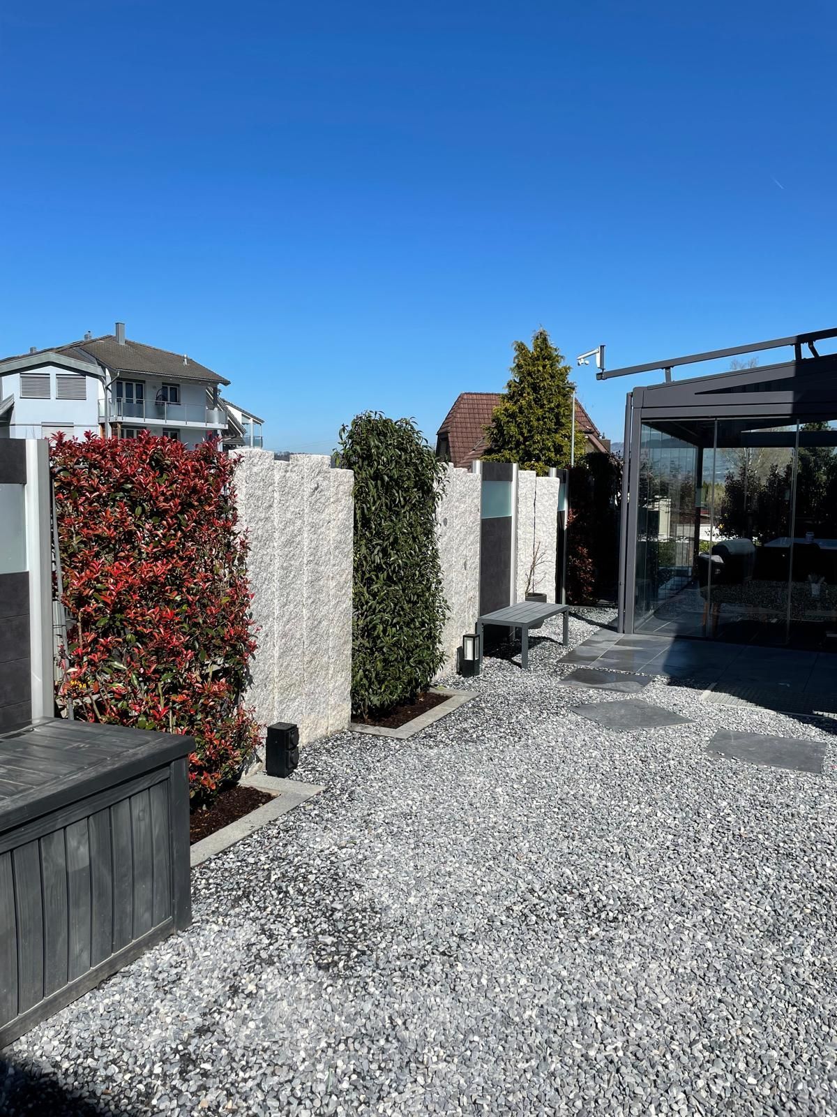 Eine Feuerstelle mit Grill, umgeben von schwarzen Steinen in einem Hof, im Hintergrund ein kleiner Teich und ein grasbewachsener Hügel.