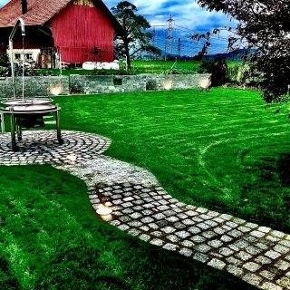 Ein Kopfsteinpflasterweg schlängelt sich durch eine grüne Rasenfläche in Richtung einer roten Scheune.