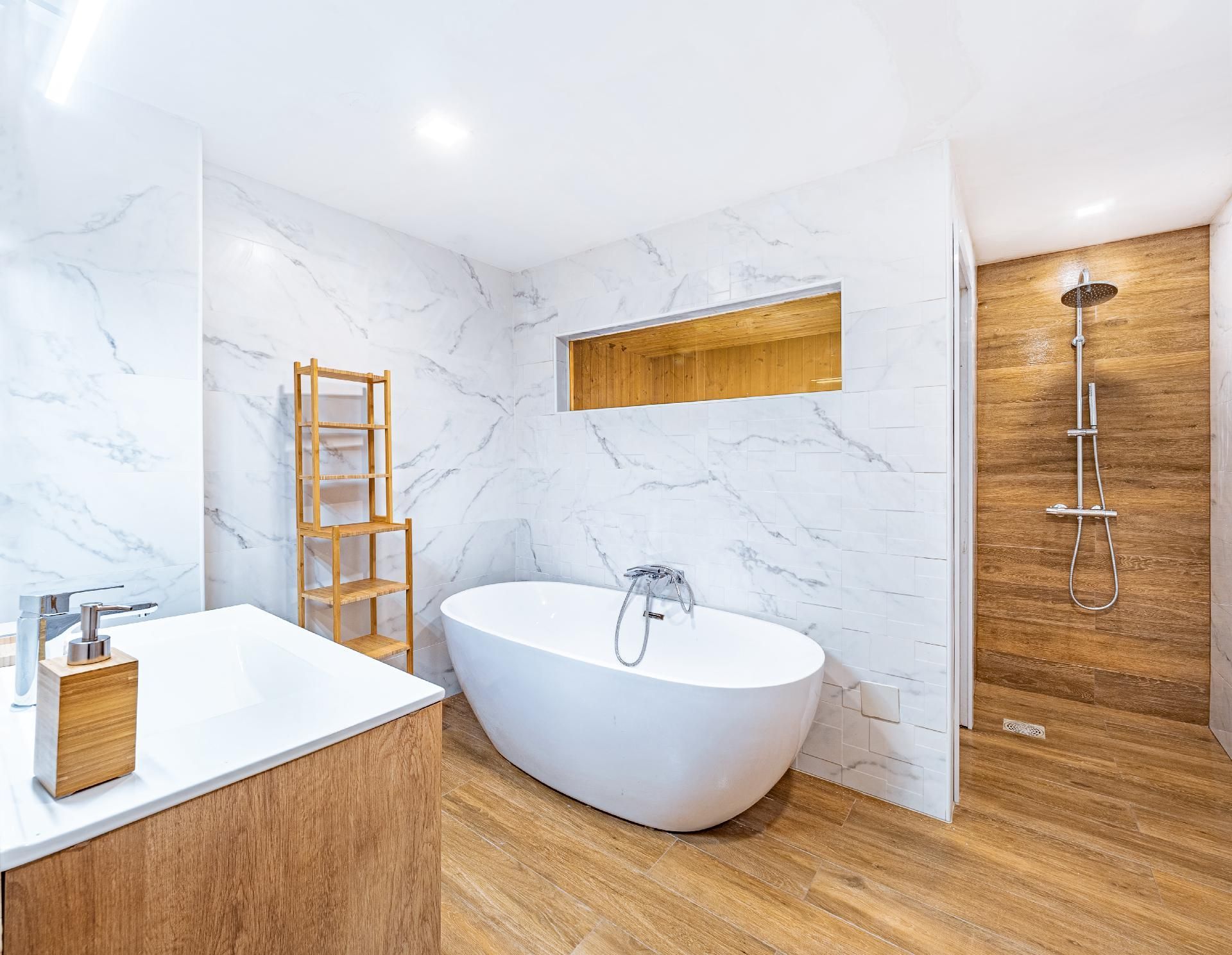 Modernes Badezimmer mit weißen Marmorwänden, Holzboden, freistehender Badewanne, Dusche und Waschtisch.