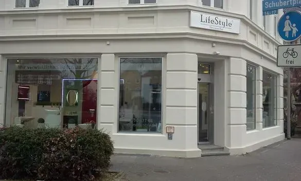 Weiße Ladenecke mit großen Schaufenstern, Boutique-Schildern und einem Fahrradweg-Schild auf dem Bürgersteig.