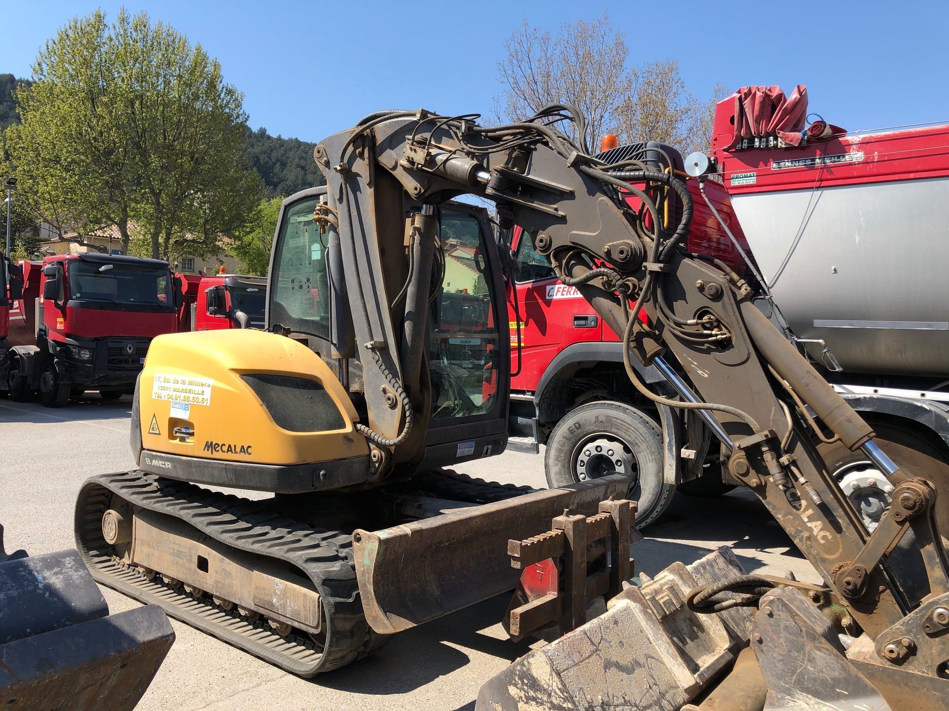 Pelle multi-fonctions Mécalac 8 à côté d'un camion rouge