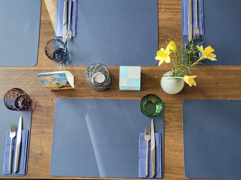 Ein Holztisch mit blauen Tischsets und Besteck und einer Vase mit Blumen.