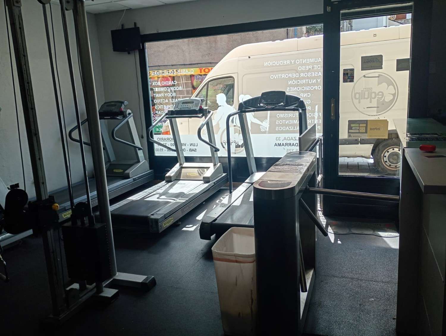 Un pequeño gimnasio cubierto con máquinas de ejercicio y una gran ventana con vistas a una furgoneta aparcada en la calle.