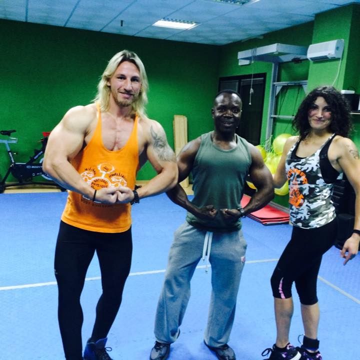 Tres personas posan en un gimnasio, flexionando los músculos de sus brazos mientras sonríen a la cámara.