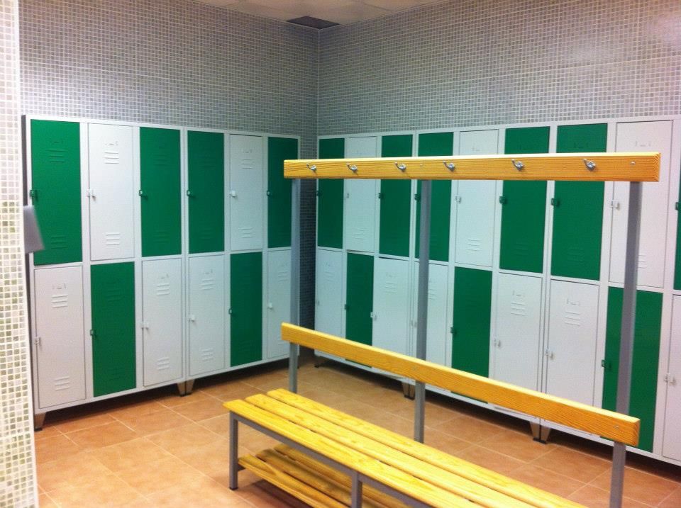 Un vestuario vacío con filas de taquillas verdes y blancas y un banco de madera en el centro.