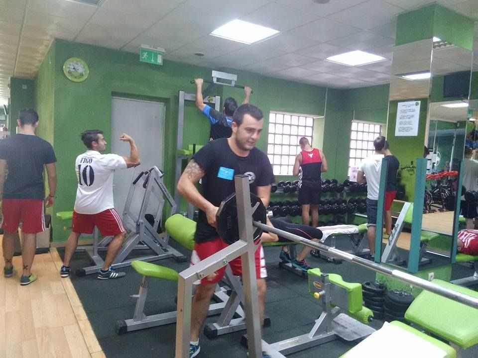 Varias personas están levantando pesas y haciendo ejercicio en un gimnasio con paredes verdes y aparatos grises.