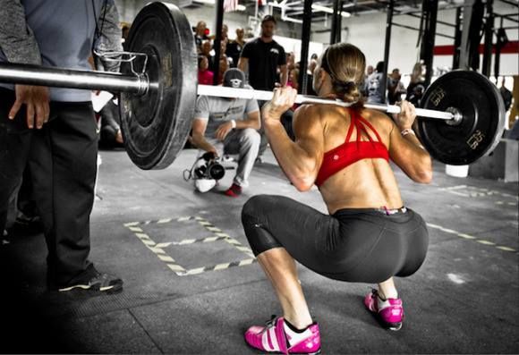 Una persona realiza una sentadilla con barra en un gimnasio, con espectadores y un fotógrafo presentes.