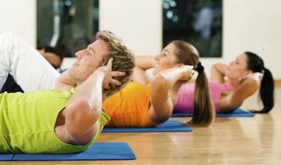 Tres personas con ropa deportiva colorida realizan abdominales sobre colchonetas en un gimnasio bien iluminado.