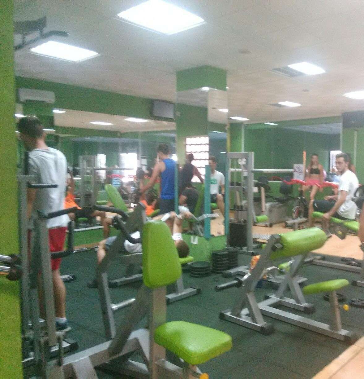 Varias personas hacen ejercicio en diversas máquinas de pesas en un gimnasio con paredes verdes y espejos.