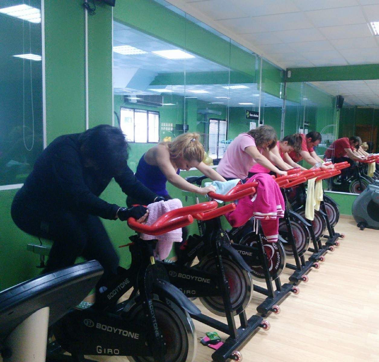 Personas en un gimnasio participando en una clase de spinning en bicicletas estáticas en una sala con espejos.