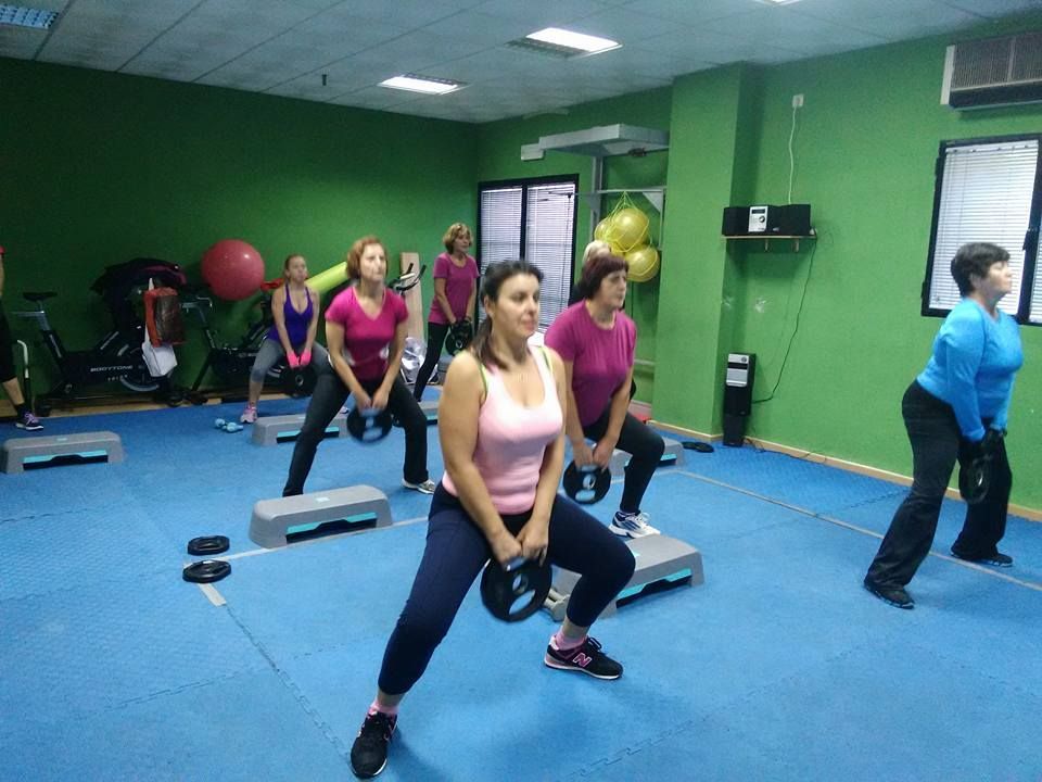 En una clase de fitness, varias personas sostienen pesas mientras realizan sentadillas en un estudio con paredes verdes.