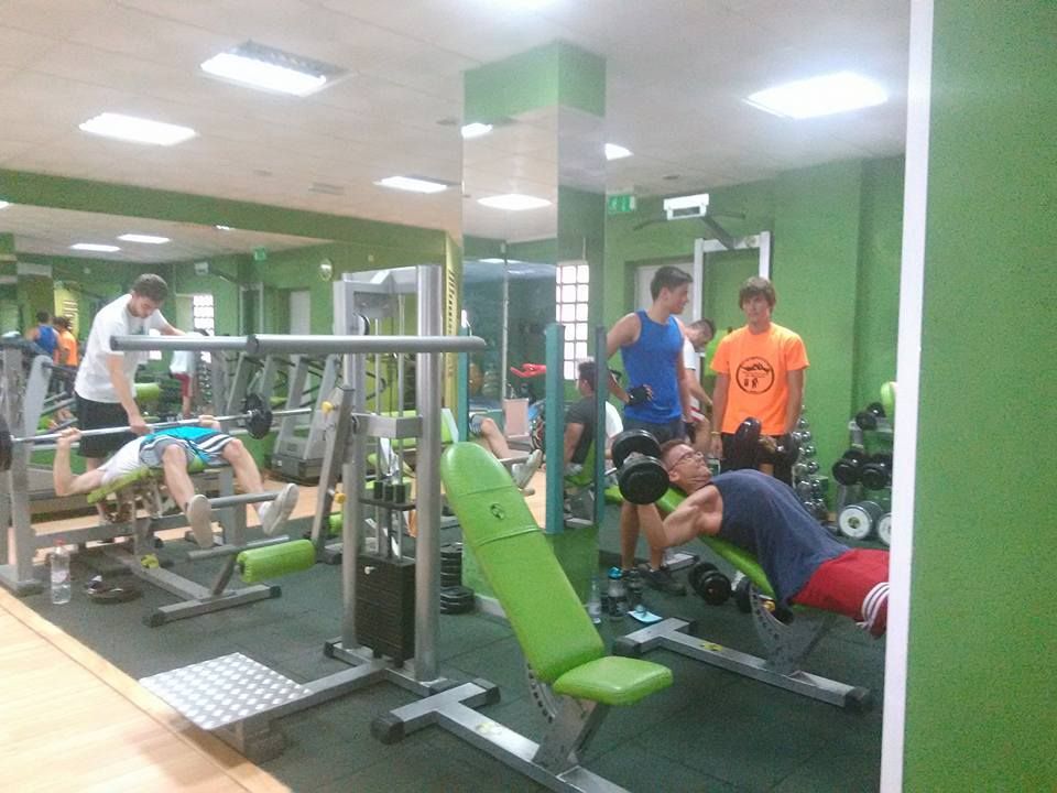 Varias personas hacen ejercicio con pesas y aparatos en un gimnasio con paredes verdes y superficies espejadas.