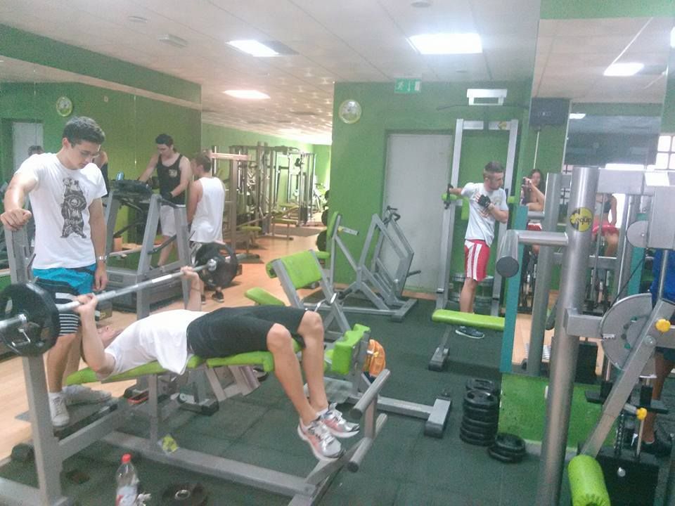 Personas haciendo ejercicio en un gimnasio con paredes verdes, bancos de pesas y máquinas.