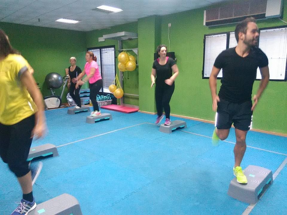 Un grupo de personas en un gimnasio haciendo ejercicio en escalones de aeróbic sobre un suelo azul frente a paredes verdes.