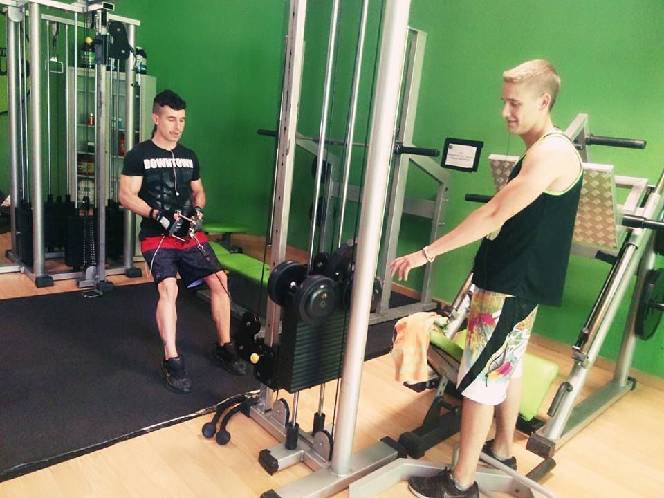Dos personas en un gimnasio: una realiza un remo con cable sentado, mientras que la otra permanece de pie cerca y observa.