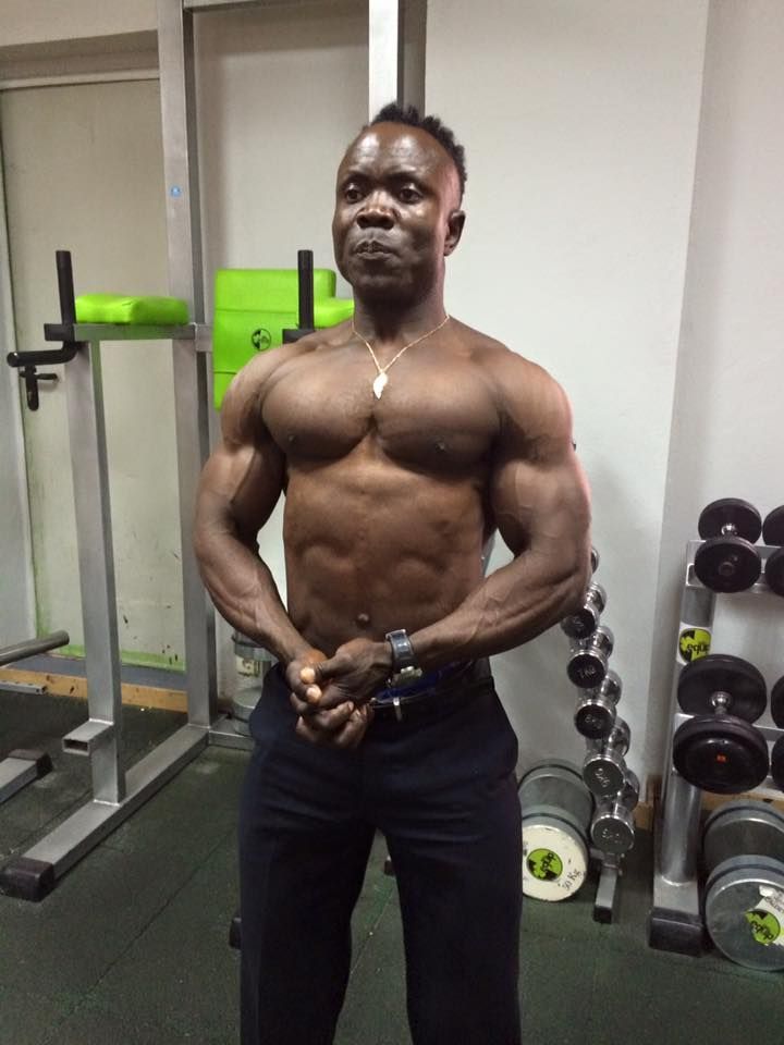 Una persona musculosa posando en un gimnasio con estantes de pesas al fondo.
