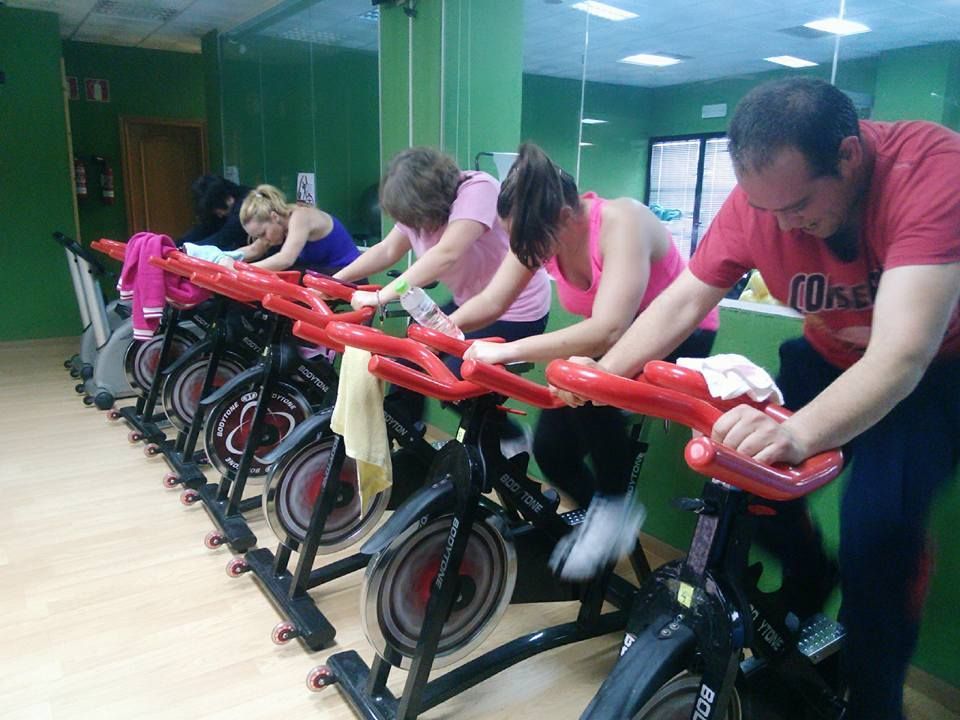 La gente hace ejercicio en bicicletas estáticas de spinning en un gimnasio con paredes verdes y suelos de madera.