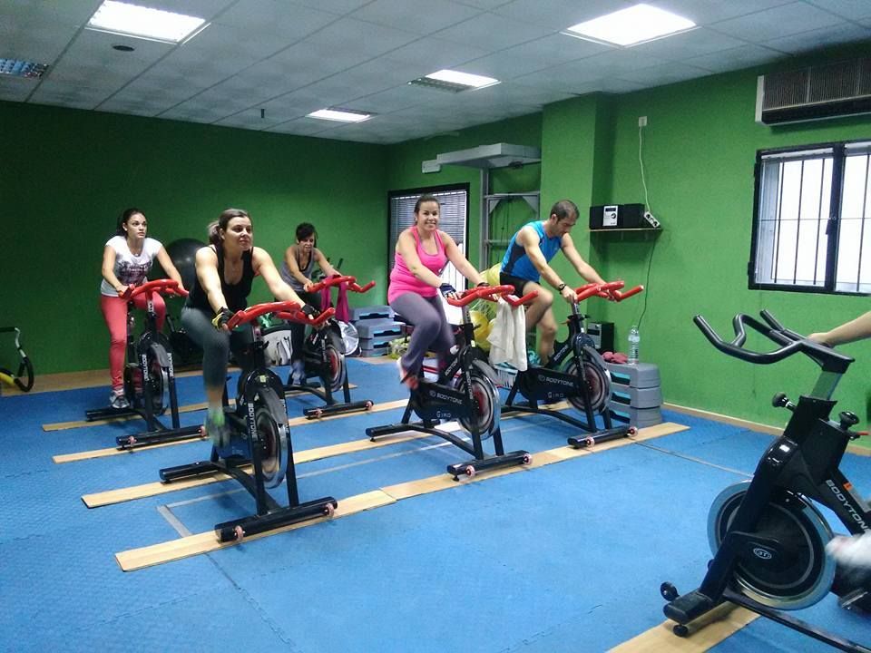 Personas haciendo ejercicio en bicicletas estáticas en un gimnasio con paredes verdes y suelo azul.