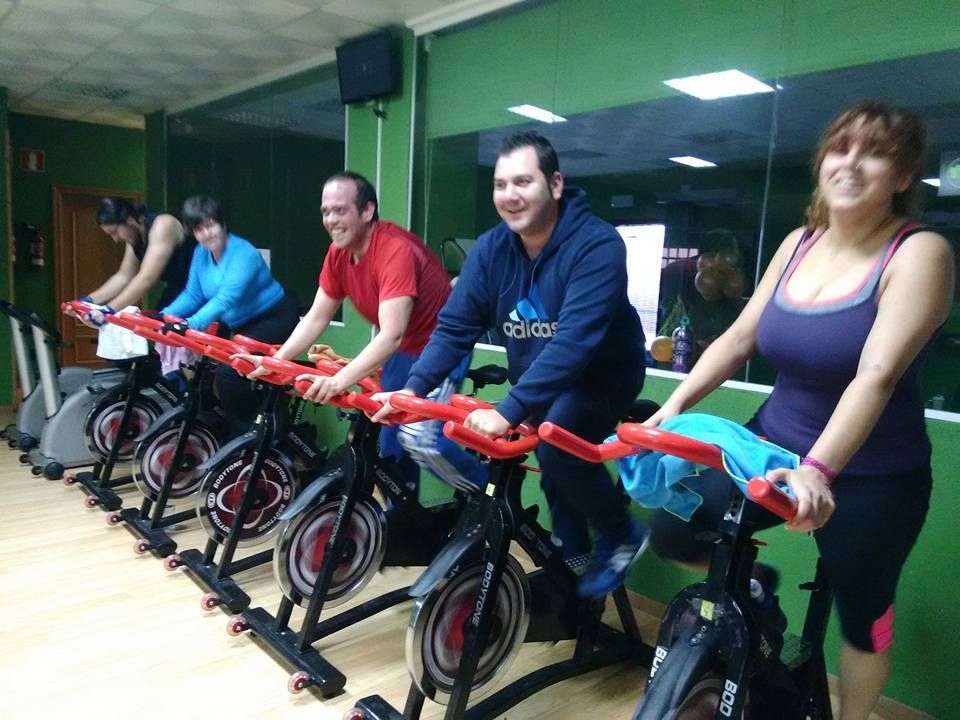 Cuatro personas sonríen mientras hacen ejercicio en bicicletas estáticas en un gimnasio con paredes verdes.