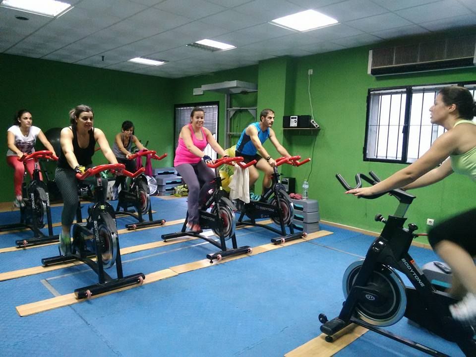 Personas haciendo ejercicio en bicicletas estáticas en un gimnasio con paredes verdes y suelo azul.