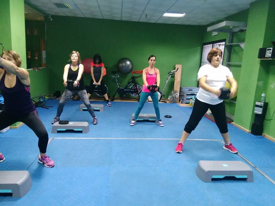 Un grupo de personas realizando sentadillas con pesas durante una clase de ejercicios en un gimnasio con paredes verdes.