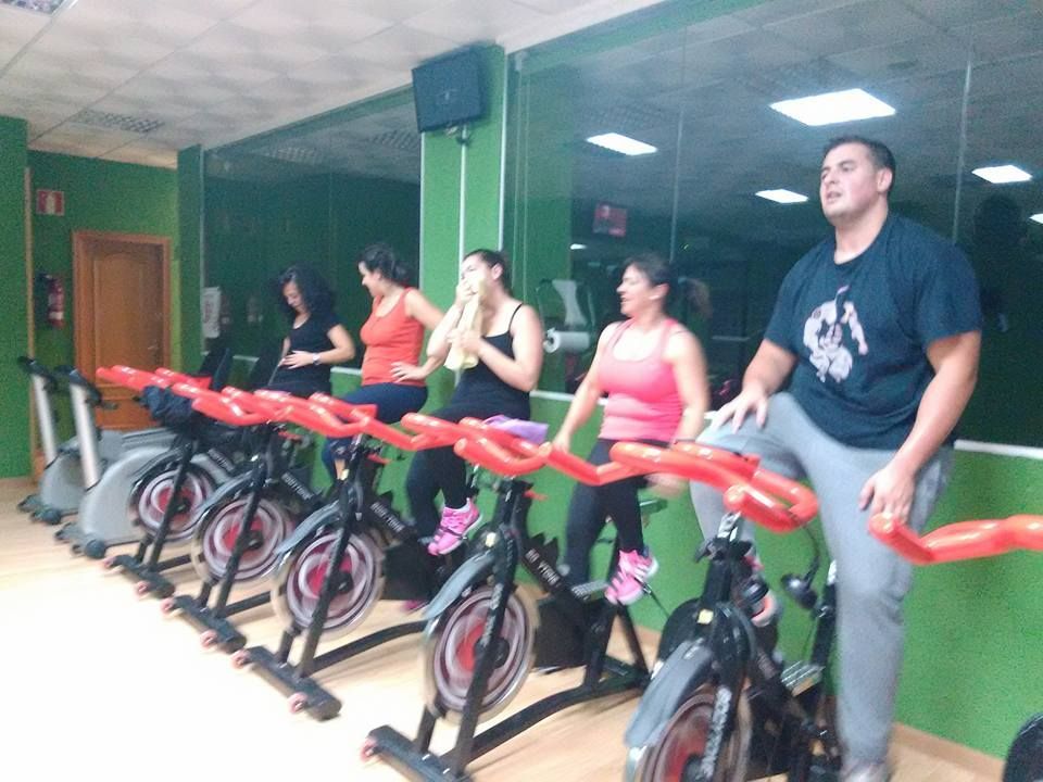 Un grupo de personas haciendo ejercicio en bicicletas estáticas en un gimnasio con paredes verdes y grandes espejos.