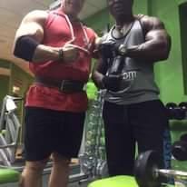 Dos hombres musculosos con ropa de gimnasio posando juntos dentro de una sala de pesas.