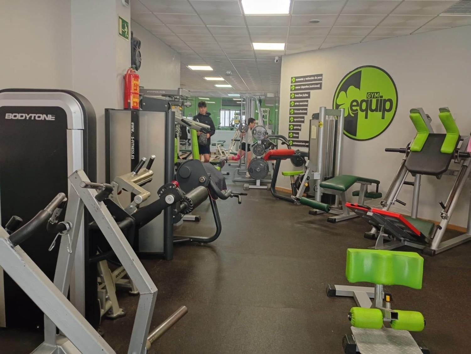 Vista interior de un gimnasio con varias máquinas de pesas, equipos de fitness y un logotipo verde en la pared.