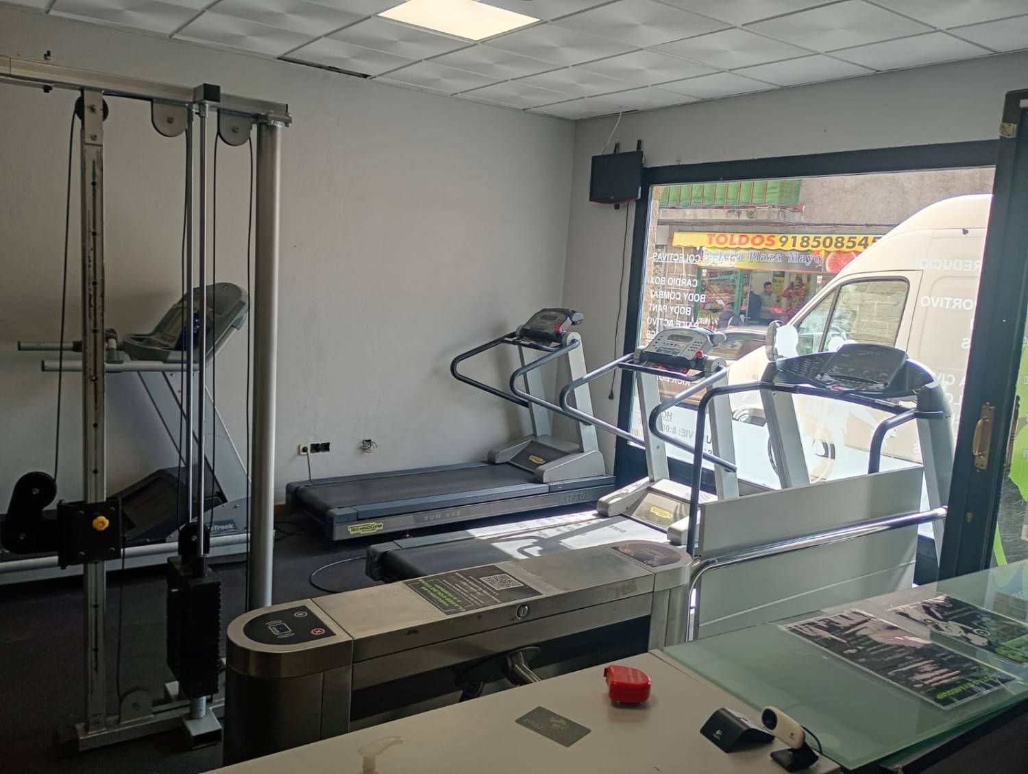 Un pequeño gimnasio con una máquina de poleas y dos cintas de correr cerca de una gran ventana con vistas a la calle.