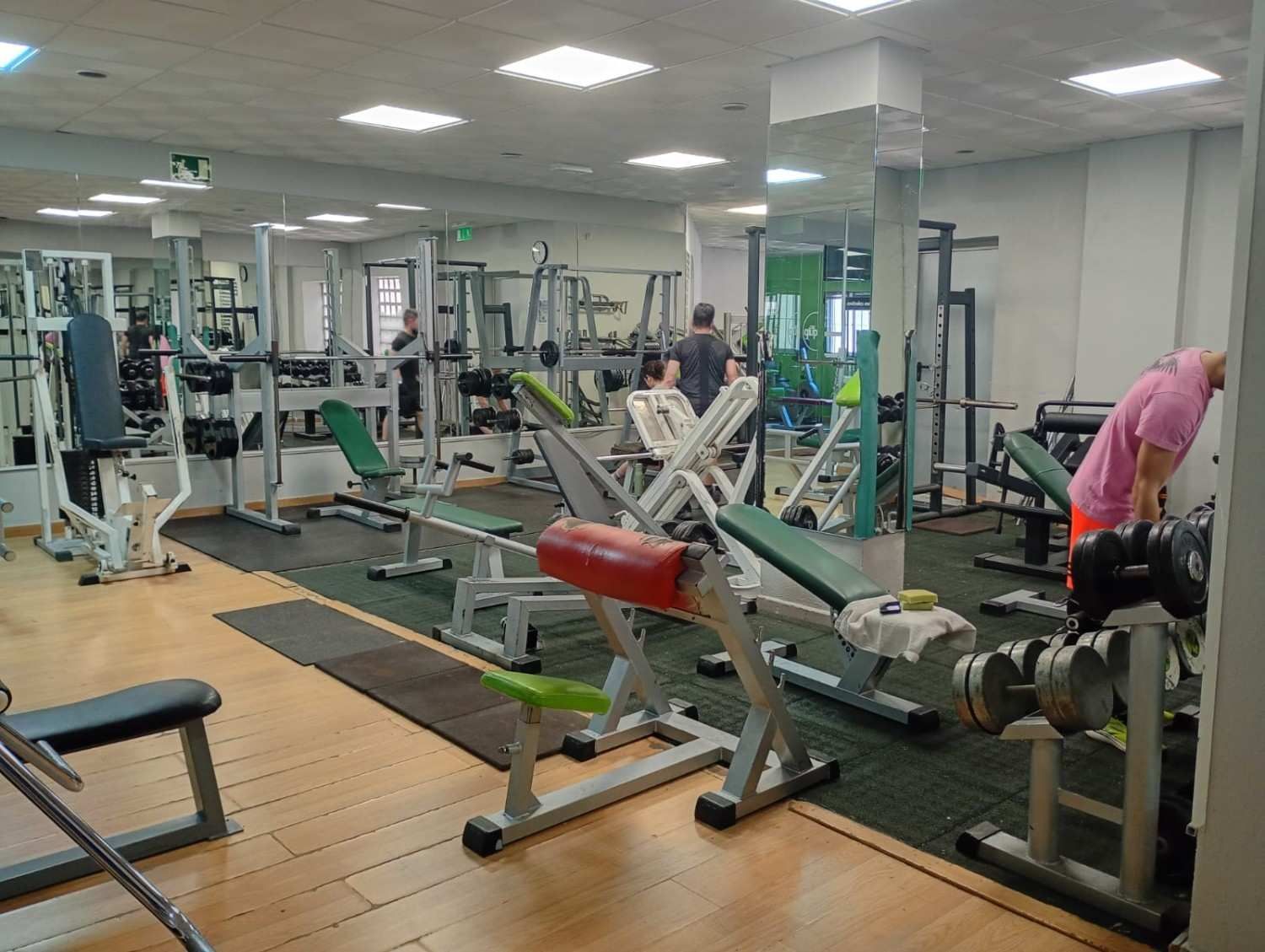 Un gimnasio bien iluminado con filas de bancos de pesas, máquinas de ejercicio y una pared con espejos que refleja el equipo.