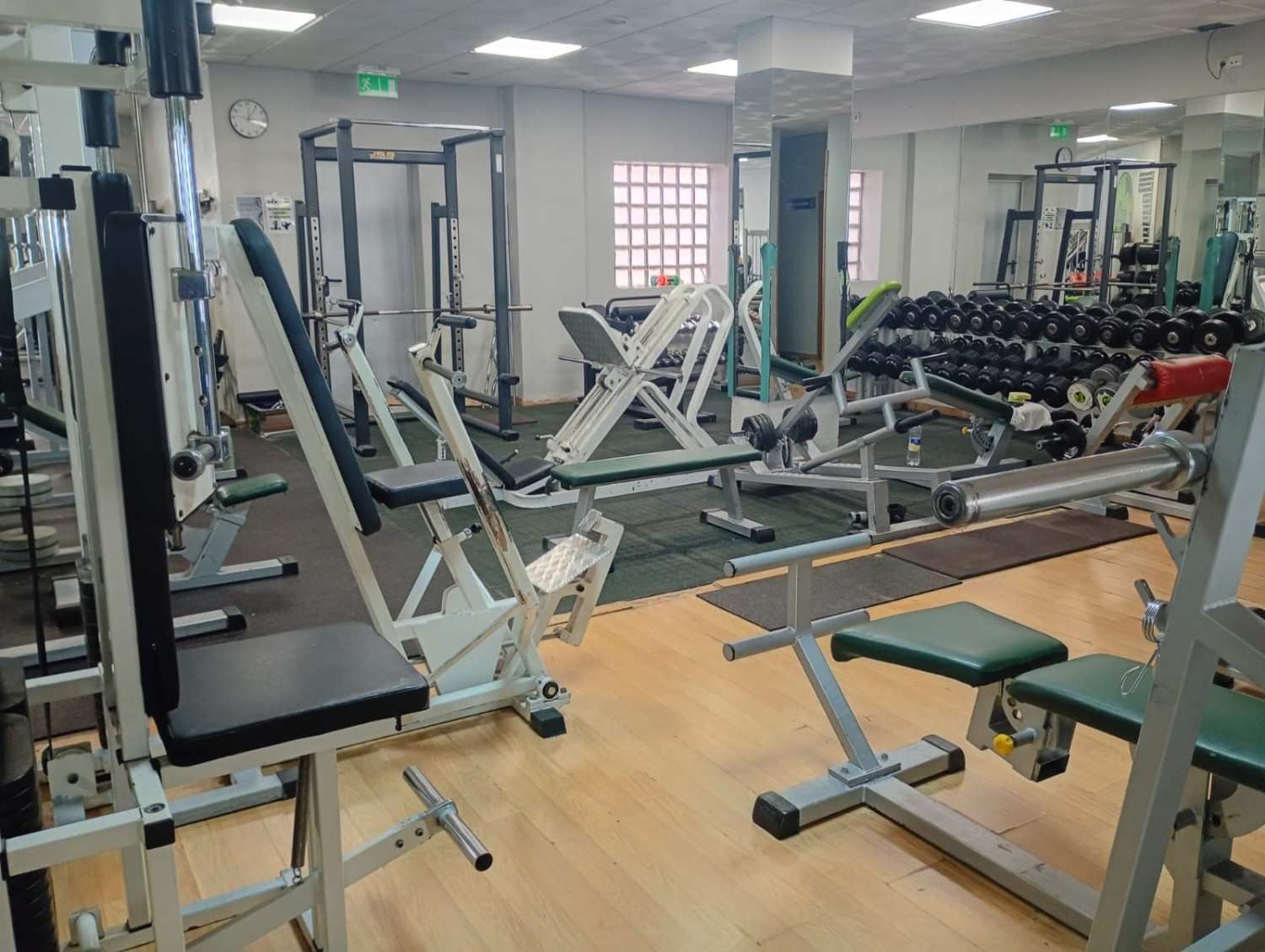 Gimnasio vacío con varias máquinas de entrenamiento de fuerza, pesas y soportes sobre un suelo de madera de color claro.