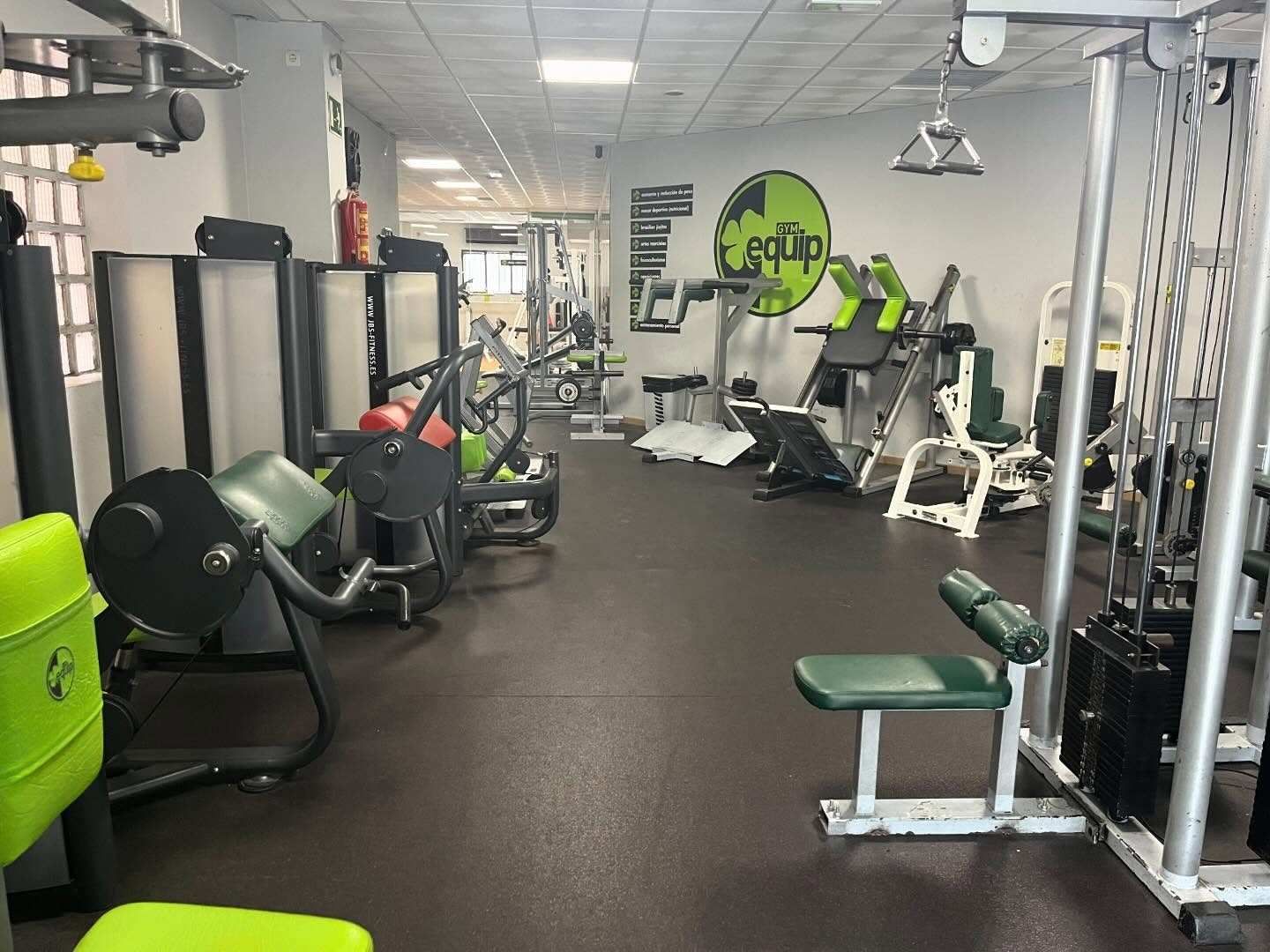 Un gimnasio pequeño y bien iluminado, con varias máquinas de musculación, suelo de goma y un logotipo verde en la pared.