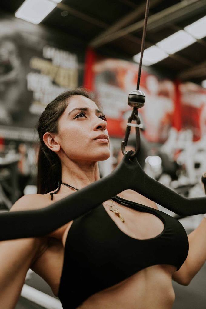 Mujer con top deportivo negro en un gimnasio, mirando hacia arriba mientras sujeta la manivela de una máquina de poleas.