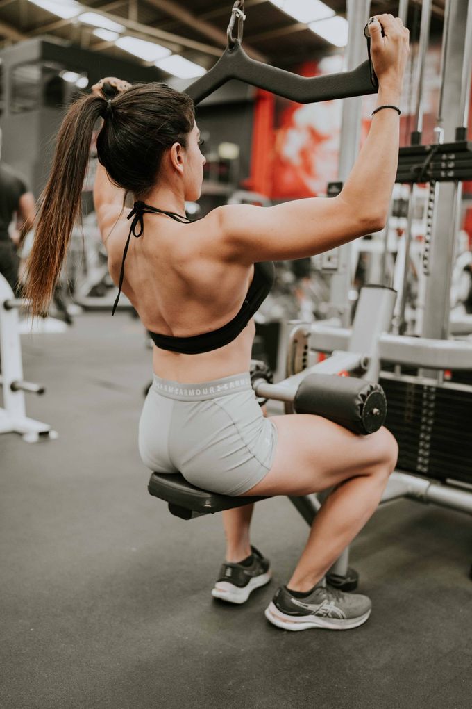 Mujer realizando un ejercicio de jalón lateral en una máquina de gimnasio, vistiendo un sujetador deportivo negro y mallas grises.