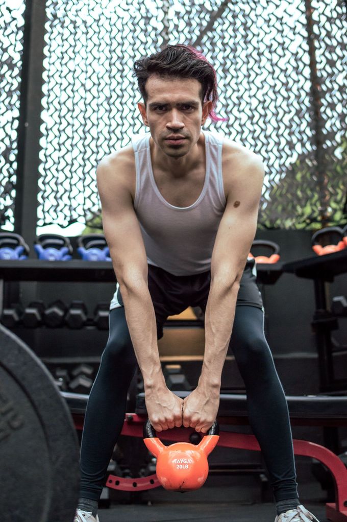 Hombre haciendo ejercicio con una pesa rusa en un gimnasio, inclinado hacia adelante, con una camiseta blanca sin mangas y mallas negras.