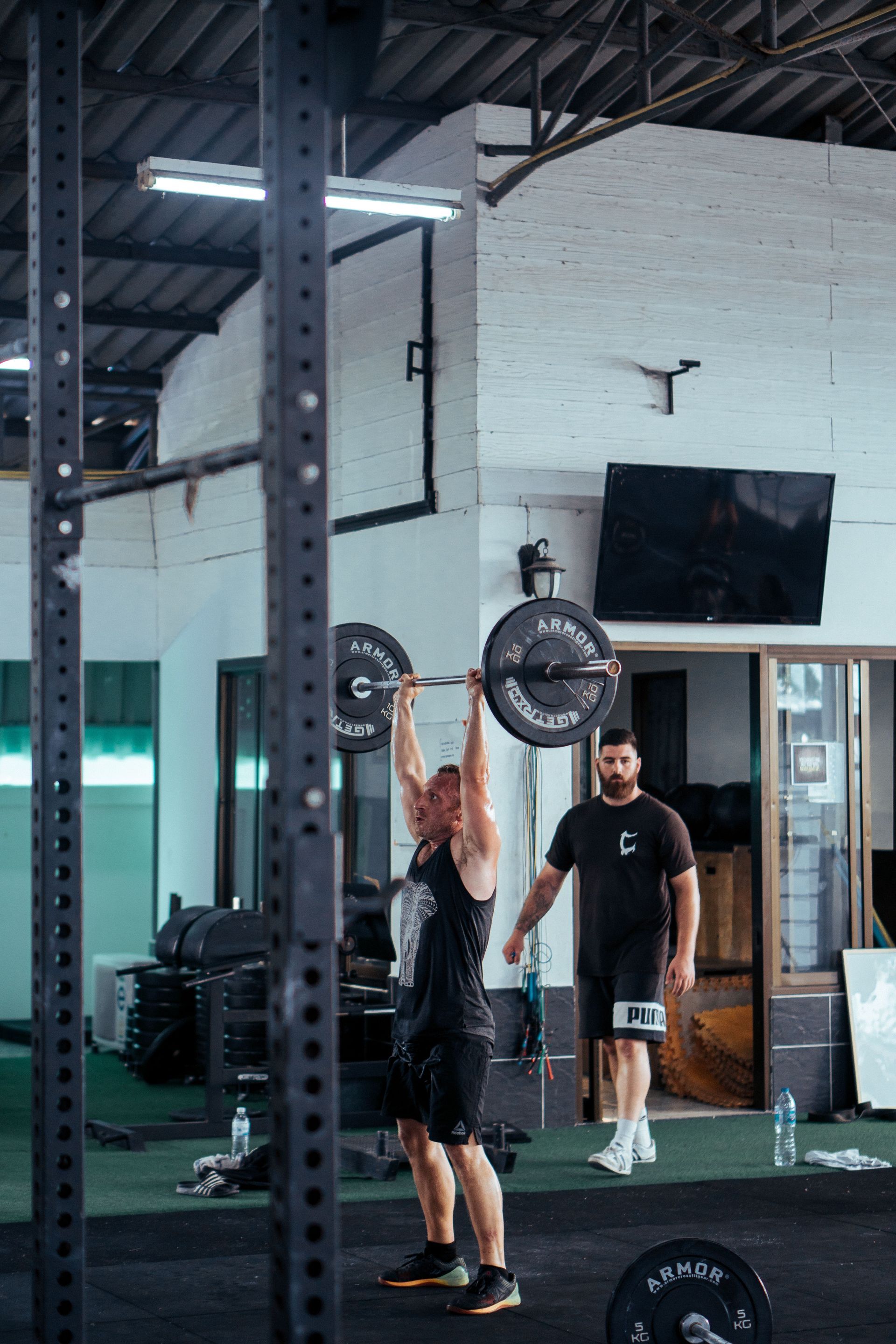 Dos personas levantando pesas en un gimnasio con techos altos y espejos.