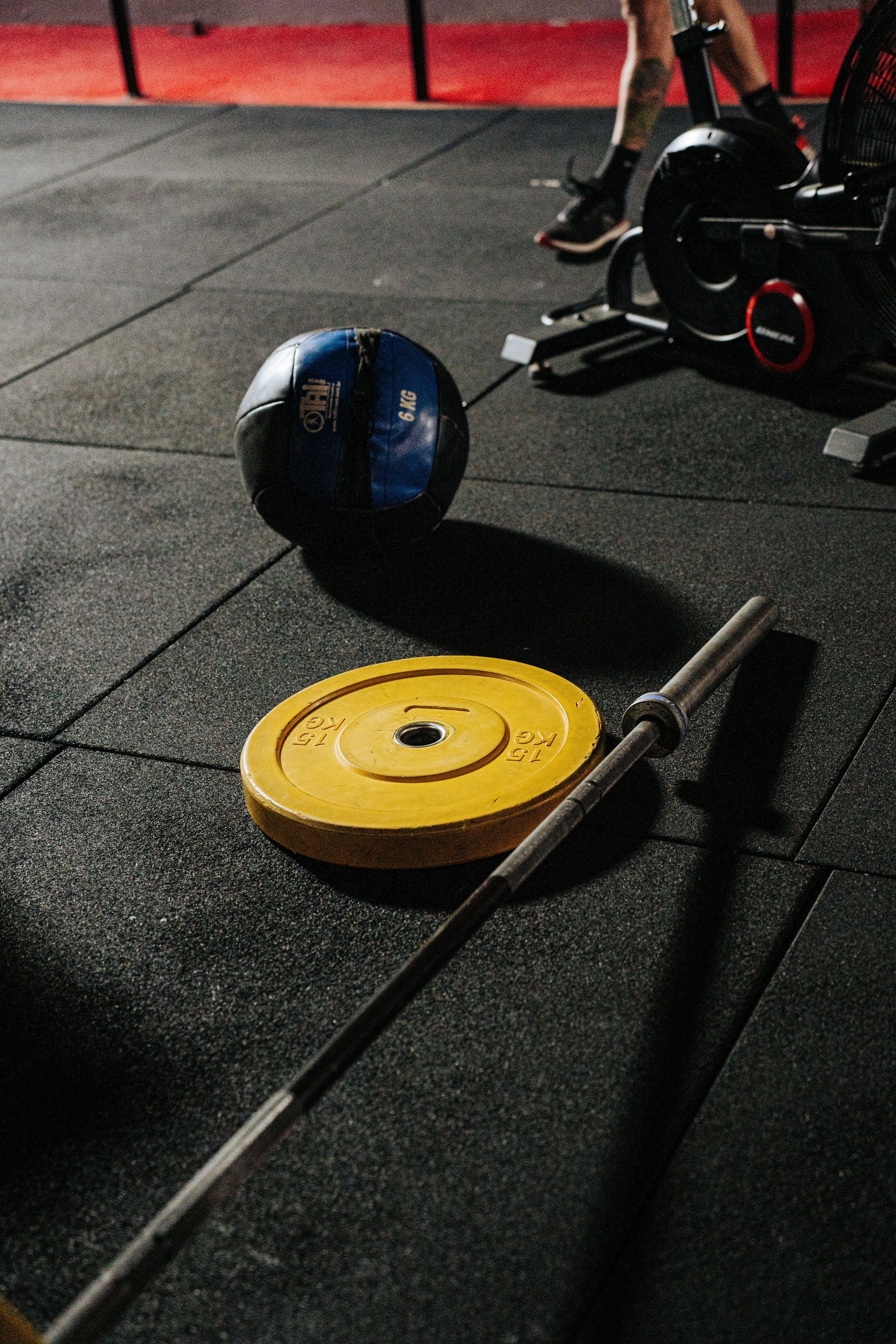 Suelo de gimnasio con una barra, un disco de pesas amarillo y una pelota medicinal cerca de las piernas de una persona.