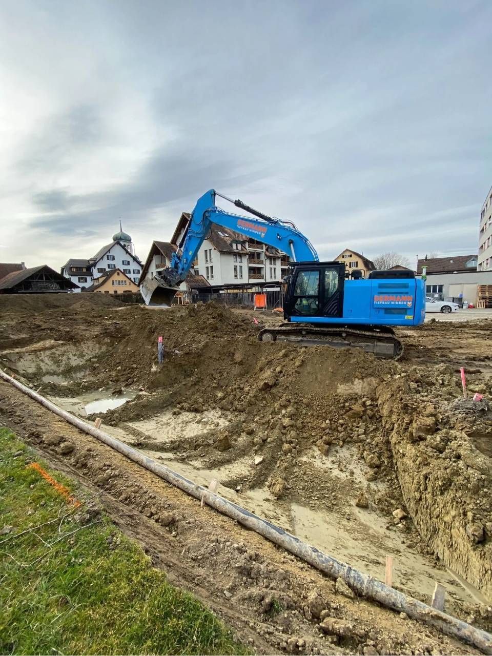 Ein blauer Bagger gräbt auf einer Baustelle in der Nähe von Gebäuden, darüber ist der Himmel bewölkt.