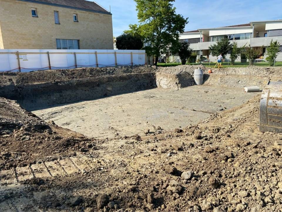 Baustelle: Ausgehobene Baugrube mit Erde, Stützmauern und Gebäuden im Hintergrund.