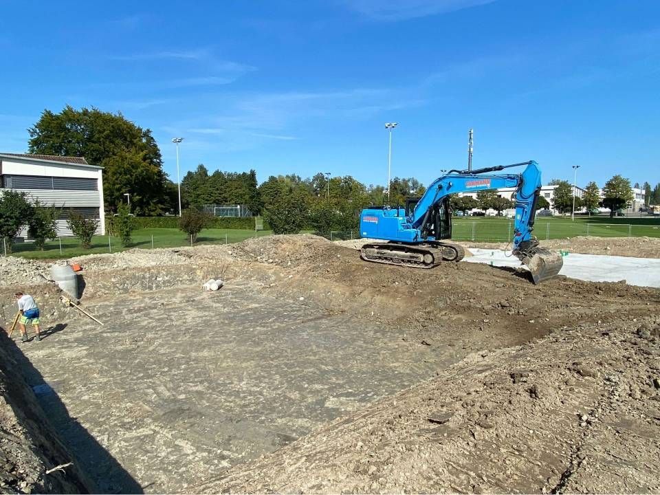 Ein blauer Bagger auf einer Baustelle, der Erde bewegt. Zwei Arbeiter stehen daneben, im Hintergrund ist klarer Himmel zu sehen.