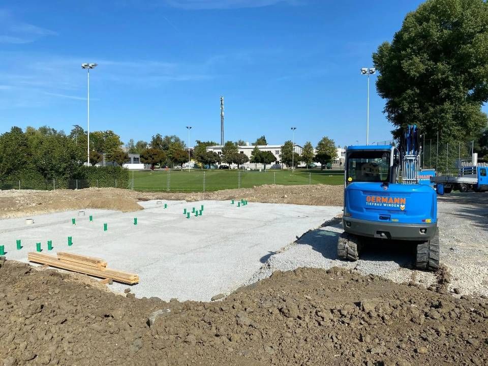 Baustelle mit blauem Bagger auf Kiesbett, grünen Pfählen, Erdhügeln und blauem Himmel.