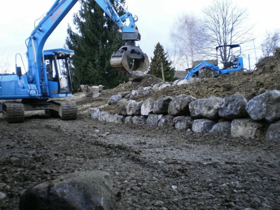 Ein blauer Bagger platziert grosse Steine ​​zum Bau einer Stützmauer im Freien.
