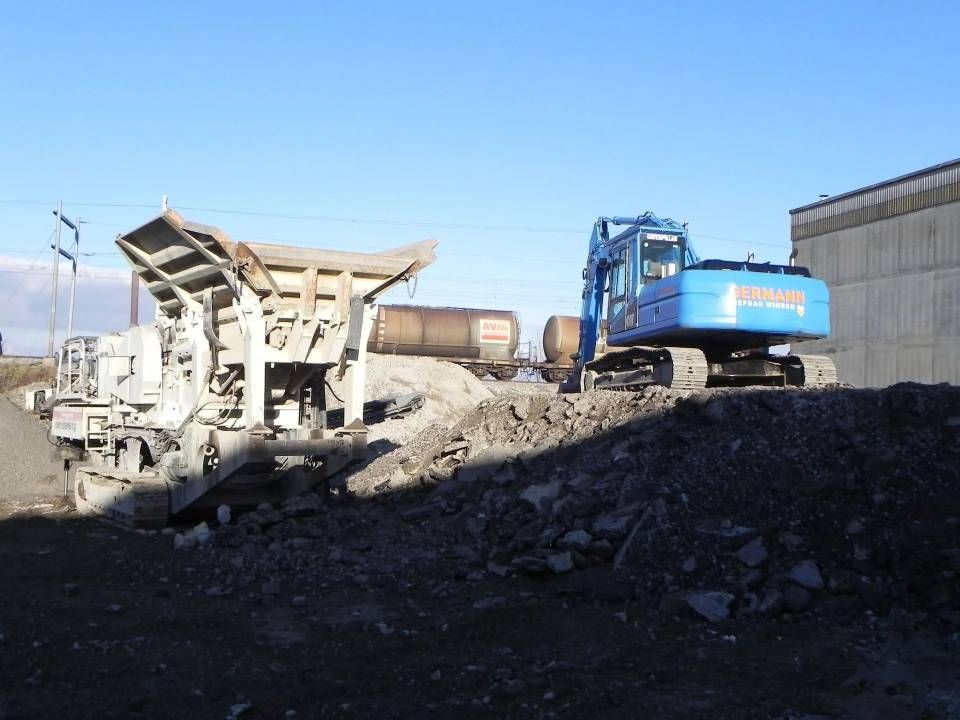 Ein blauer Bagger und ein Betonbrecher auf einem Schutthaufen vor klarem Himmel.