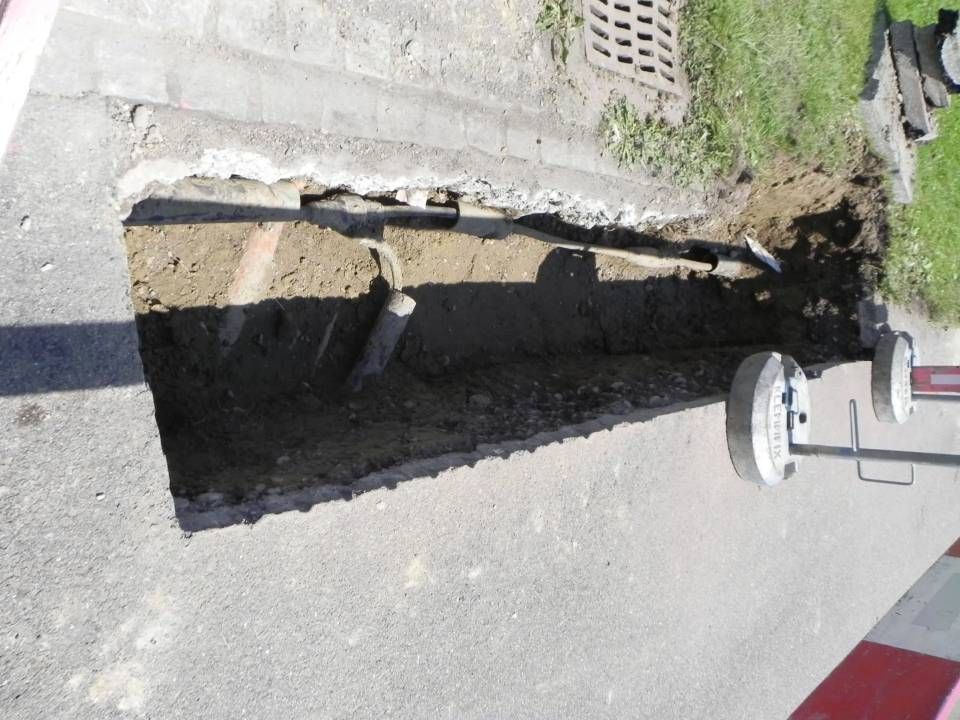 Offener Graben im Asphalt neben einem Bordstein und einer Rasenfläche. Betonblöcke und Rohre sind im Graben sichtbar.
