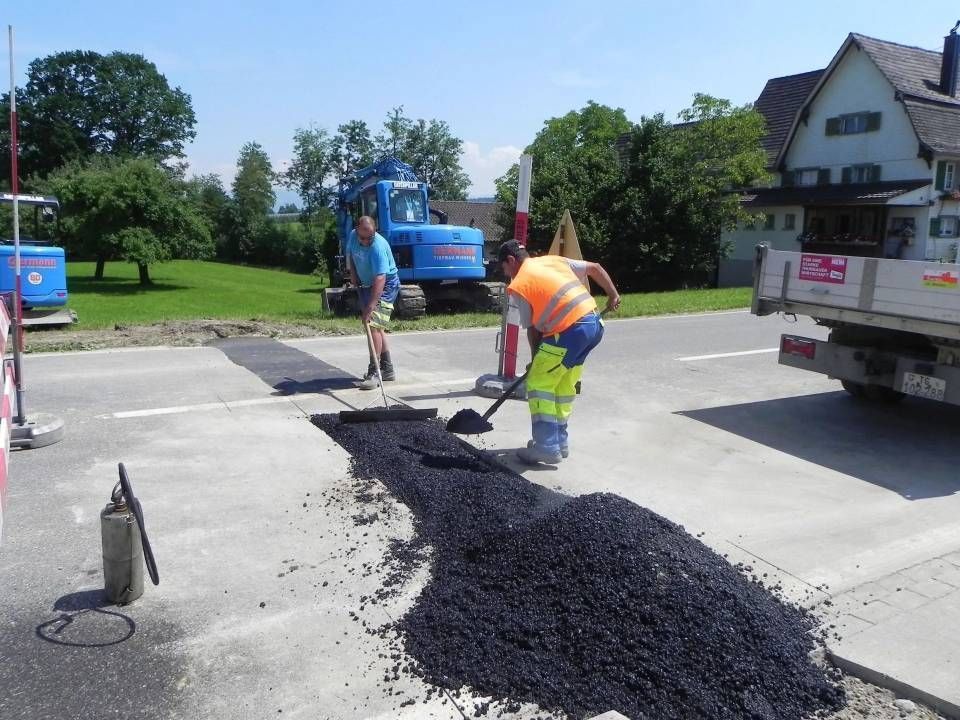 Strassenarbeiter asphaltieren eine Strasse. Einer schaufelt Asphalt, ein anderer steht daneben; Lastwagen und Bagger sind zu sehen.