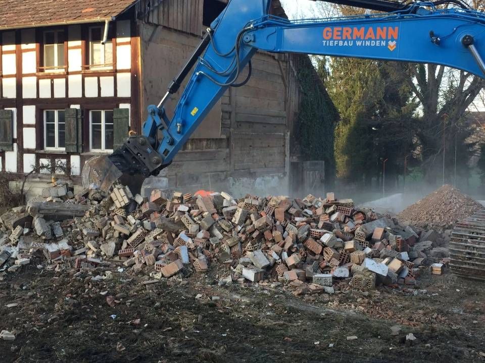 Ein Bagger reisst ein Backsteingebäude ab; im Hintergrund ist ein Holzgebäude zu sehen.