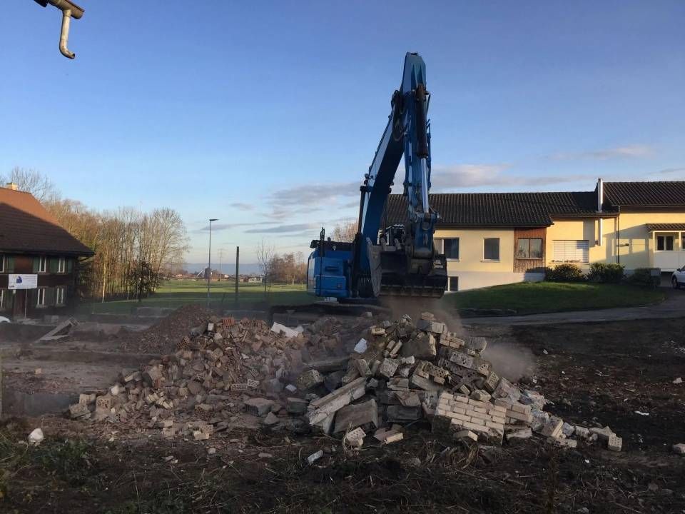 Ein blauer Bagger reisst an einem sonnigen Tag einen Haufen Betonschutt neben Gebäuden ab.