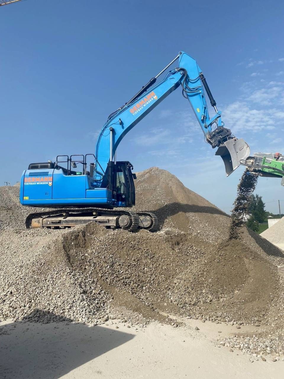 Ein blauer Bagger verlädt unter klarem Himmel Kies auf ein grünes Förderband.