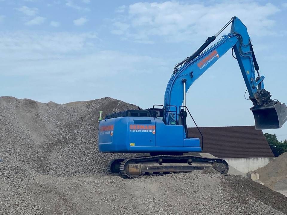 Blauer Bagger mit angehobenem Arm und Schaufel, der Kies auf einer Baustelle bewegt.